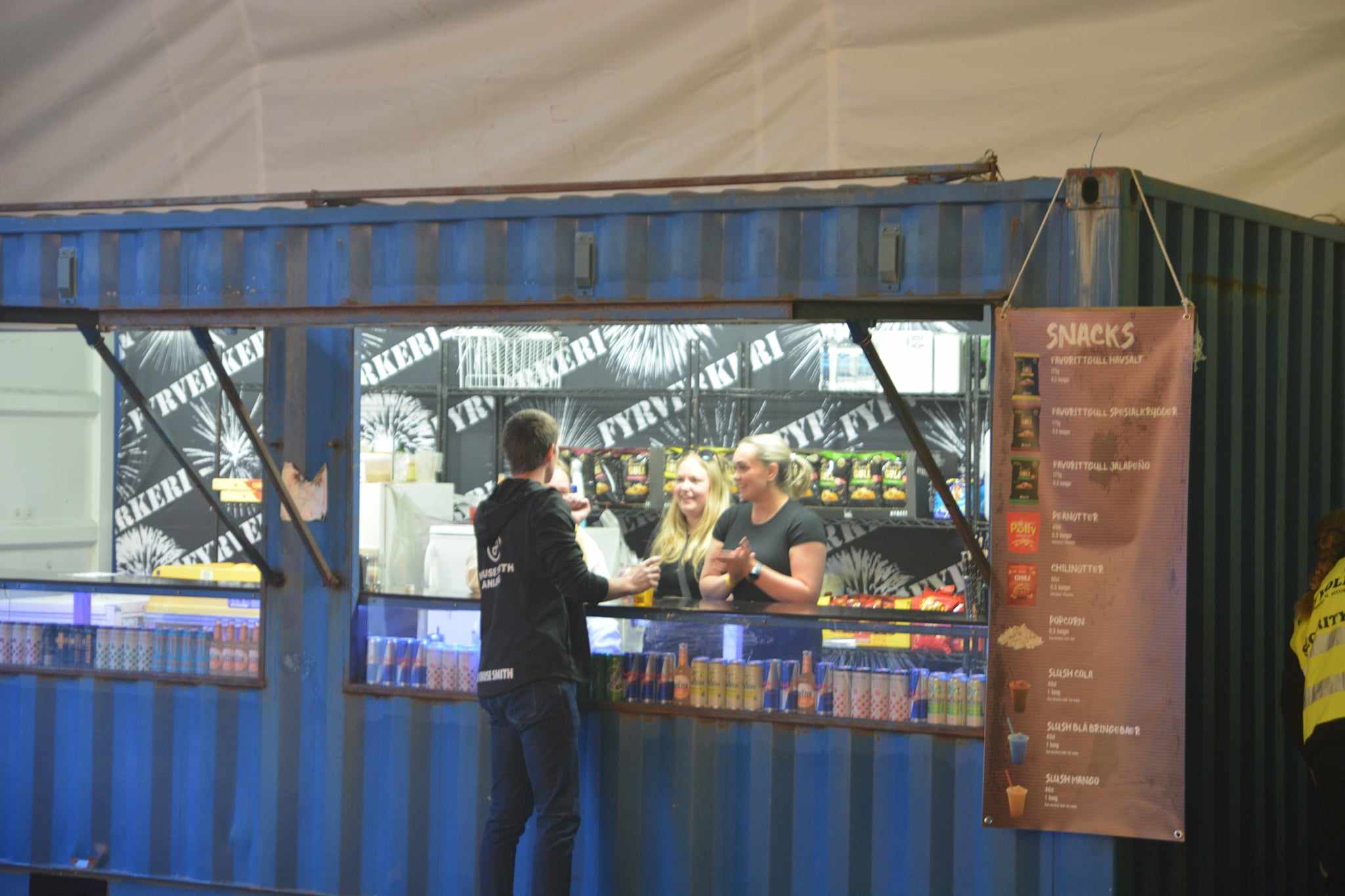 En bar og snackdisk inne i en blå fraktkonteiner, med ulike drikkevarer og matvarer utstilt, og kunder i samhandling med personalet.